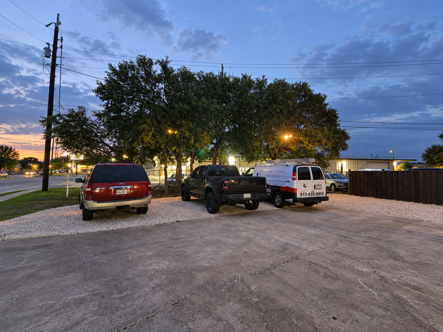 Three vehicles parked in a commercial lot at dusk, with exterior lighting illuminating mature trees and the surrounding area.