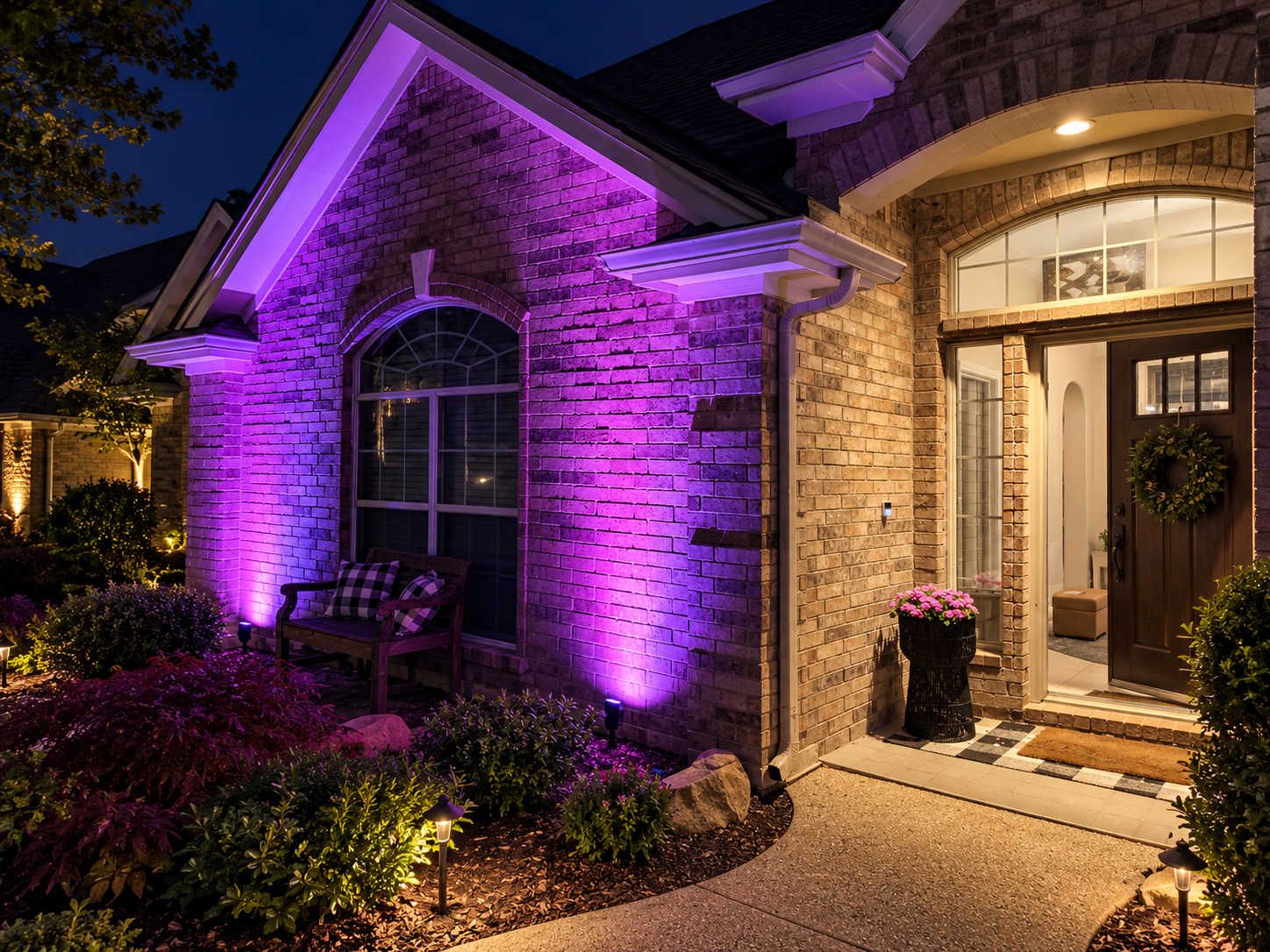 A brick home features vibrant purple and magenta exterior lighting, a warmly lit entryway, and path lights along the curved walkway.