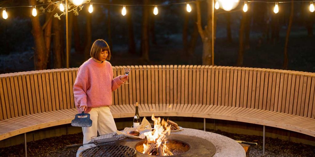 A woman in a pink sweater holds a wine glass near a glowing fire pit surrounded by curved wooden seating.